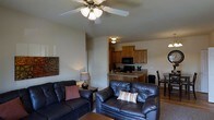 This single-floor space offers a well-designed open-concept layout that seamlessly connects the living area and kitchen, creating a welcoming and functional environment. The living room is anchored by two dark leather sofas arranged around a coffee table, providing a comfortable and inviting seating arrangement. A ceiling fan with a light fixture overhead ensures both comfort and ambient lighting throughout the space. Warm neutral-toned walls complement the rich tones of the furniture and flooring, contributing to the overall cozy atmosphere. Decorative wall art adds a touch of character to the room. The kitchen is outfitted with warm honey-toned wood cabinetry that extends to the upper walls, offering generous storage. Black appliances, including a microwave and range, provide a sleek contrast to the cabinetry. A peninsula-style breakfast bar with a countertop overhang serves as both a food preparation area and an informal dining spot. Adjacent to the kitchen, a dining area with a table and chairs provides a dedicated space for meals. The space benefits from a practical, efficient floor plan that makes excellent use of every area, making it well-suited for everyday living.
