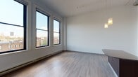 This thoughtfully designed single-floor urban residence blends contemporary finishes with an industrial-inspired aesthetic. The open-concept living and kitchen area commands attention with its dramatically high ceilings, exposed ductwork, and light hardwood flooring that extends throughout the main living spaces. Pendant lighting hangs above a granite-topped kitchen island, complementing the full suite of stainless steel appliances — including a refrigerator, dishwasher, and range — all set against rich dark wood cabinetry with granite countertops and a double sink.

Large, black-framed windows flood the living areas with natural light while offering sweeping views of the surrounding urban landscape, including rooftops and rolling hills in the distance. The bedrooms feature plush carpeting and their own generous windows, maintaining the unit's connection to the outdoors. A dedicated laundry area with a washer and dryer adds everyday convenience, while the bathroom is equipped with an accessible-style tub and handrails. The overall ambiance is bright, open, and modern, with clean white walls and an efficient layout that makes excellent use of every area of the space.