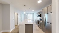 This thoughtfully designed one-bedroom, one-bathroom residence in Baton Rouge, Louisiana, offers a modern and cohesive aesthetic across its efficiently utilized single-floor layout. The open-concept kitchen serves as a central highlight, boasting granite countertops, a large center island with pendant lighting, gray shaker-style cabinetry extending to the ceiling, a white subway tile backsplash, and a full suite of stainless steel appliances. Light wood-style flooring flows throughout the main living areas, enhancing the bright, airy atmosphere created by neutral wall tones and recessed lighting. The living and dining area is generously proportioned, featuring a ceiling fan and a set of French doors that invite natural light and provide access to the outdoors. The spacious primary bedroom is finished with plush carpet and includes a large walk-in closet with built-in wire shelving for ample storage. The bathroom continues the refined design language with gray cabinetry, granite countertops, and a tub-shower combination with tile surround. Clean lines, consistent finishes, and a well-considered floor plan make this residence a functional and attractive living space.
