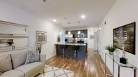 This contemporary single-floor space is thoughtfully designed with a seamless open-concept layout that connects the living and kitchen areas into one cohesive, flowing environment. Warm-toned hardwood flooring runs throughout, anchoring the space with a sense of continuity and warmth.

The kitchen is a standout feature, outfitted with sleek dark cabinetry, stainless steel appliances including a refrigerator with ice maker, microwave, and range, as well as a large central island with a built-in sink and bar-height seating for two. Three pendant lights hang above the island, providing focused task lighting that also serves as a modern design accent. Recessed lighting across the ceiling ensures the entire space is bright and well-illuminated.

The living area is open and inviting, complemented by clean white walls that enhance the sense of space and light. A patterned area rug and a light-toned sofa add comfort and style, while decorative greenery brings a natural element into the interior. Wall art and thoughtful décor details contribute to the space's polished, modern aesthetic.

A hallway visible from the main area suggests additional rooms beyond, adding to the overall functionality of this well-appointed space.