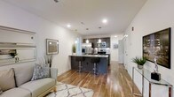 This contemporary single-floor space is thoughtfully designed with a seamless open-concept layout that connects the living and kitchen areas into one cohesive, flowing environment. Warm-toned hardwood flooring runs throughout, anchoring the space with a sense of continuity and warmth.

The kitchen is a standout feature, outfitted with sleek dark cabinetry, stainless steel appliances including a refrigerator with ice maker, microwave, and range, as well as a large central island with a built-in sink and bar-height seating for two. Three pendant lights hang above the island, providing focused task lighting that also serves as a modern design accent. Recessed lighting across the ceiling ensures the entire space is bright and well-illuminated.

The living area is open and inviting, complemented by clean white walls that enhance the sense of space and light. A patterned area rug and a light-toned sofa add comfort and style, while decorative greenery brings a natural element into the interior. Wall art and thoughtful décor details contribute to the space's polished, modern aesthetic.

A hallway visible from the main area suggests additional rooms beyond, adding to the overall functionality of this well-appointed space.