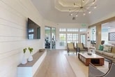 Located at 9800 Touchton Rd in Jacksonville, FL, this single-floor community clubhouse offers a thoughtfully designed gathering space that blends modern farmhouse aesthetics with resort-style comfort. The main lounge area is anchored by a striking tray ceiling with an elegant sputnik chandelier, shiplap accent walls, and light wood-look flooring that flows throughout. A wall-mounted flat-screen TV is paired with a floating shelf, while a lush vertical garden installation adds a vibrant natural element to the interior. Large multi-pane windows and glass doors flood the space with abundant natural light and offer views of the surrounding outdoor amenities, including a pool area. An adjacent game and gathering room features soaring ceilings with dramatic arched windows, pendant lighting, a high-top communal table, and a wall-mounted oversized Scrabble board, creating an engaging social setting. The well-appointed kitchen includes white shaker cabinetry with brass hardware, a shiplap-paneled island with bar seating, stainless steel appliances, a textured herringbone tile backsplash, and globe pendant lights. A separate lounge nook with modern accent chairs and patterned carpet tile provides a quieter retreat. Sliding glass doors lead to an on-site fitness center, rounding out this versatile and inviting community space.