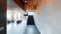 This sophisticated two-floor amenity space masterfully blends industrial and contemporary design elements throughout. Exposed concrete columns, raw concrete ceilings, and warm wood paneling create a compelling visual contrast, while dramatic Edison-bulb installations suspended on black pipe frameworks serve as striking focal points across both levels.

The lower floor anchors the space with an expansive open lounge and social area. A comfortable seating arrangement with plush sofas surrounds a central gathering zone, adjacent to a substantial communal dining table with modern white chairs and a built-in fireplace set against a textured concrete wall. A wet bar and kitchenette features sleek countertops, a sink, under-counter refrigeration, and a bank of wall-mounted flat-screen televisions, making it ideal for entertaining.

A glass-railed staircase connects the two levels, offering panoramic floor-to-ceiling views of the surrounding urban skyline that flood both floors with natural light. The upper floor provides additional lounge and recreation space, including a pool table and another wet bar station with multiple mounted screens, all framed by the same expansive glazed curtain wall. Rich hardwood flooring flows throughout, unifying the space and complementing the raw, industrial architectural palette.