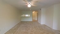 This single-floor space offers an open-concept living area finished with wall-to-wall neutral carpeting that creates a clean, understated foundation throughout. A white ceiling fan with an integrated light fixture serves as a functional centerpiece overhead, contributing to both air circulation and ambient lighting in the main living area.

A defining architectural feature is the pass-through window connecting the kitchen to the living area, allowing visibility between the two spaces and enhancing the overall sense of openness. The kitchen, visible through the pass-through, benefits from its own overhead lighting and appears to include cabinetry with white finishes. A small ledge along the pass-through offers a convenient surface for casual use.

The walls are painted in a soft, neutral tone that complements the light carpeting and contributes to a bright, airy feel. A hallway visible toward the rear of the space leads to additional rooms, indicating a practical layout with clear circulation. A wall-mounted HVAC vent is also visible, suggesting climate control throughout. Overall, the space presents a simple, functional design with a cohesive neutral palette.