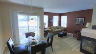 This single-floor space combines a dining area and living room in an open-concept layout that feels both functional and inviting. The dining area is anchored by a round glass-top table surrounded by dark leather chairs, with a classic pendant light hanging overhead. A large sliding glass door floods this zone with natural light and provides direct access to an outdoor deck, offering pleasant outdoor views of greenery beyond.

The living room features a bold, deep reddish-brown accent wall that creates a warm, cozy atmosphere, contrasted by lighter neutral tones on the remaining walls and ceiling. Plush wall-to-wall carpeting runs throughout, contributing to the comfortable feel of the space. A generously sized tan sofa with decorative pillows serves as the focal point of the seating area, complemented by round ottoman-style coffee tables and side tables.

Multiple windows along the living room walls allow for additional natural light, enhancing the bright and airy quality of the space. Decorative touches, including a round wall clock and tabletop accents, add character throughout. The overall ambiance is warm, casual, and well-lit, making the space feel welcoming and livable.