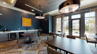 Located at 309 Mirroton Ave in Lakeland, Florida, this single-floor community amenity space combines contemporary design with a warm, inviting atmosphere. The space is divided into distinct yet cohesive areas, each thoughtfully appointed with high-end finishes and bold design choices.

The lounge area makes an immediate impression with its deep charcoal textured accent wall, a large-scale pendant light with a gold interior, and a rich cognac leather sectional sofa anchored by a geometric patterned area rug. Accent chairs in white and gold complement the curated arrangement, while a wall-mounted flat-screen TV and colorful abstract artwork add personality. Large floor-to-ceiling windows flood the space with natural light and offer views of the surrounding landscape.

The dining and gathering area continues the sophisticated aesthetic with deep navy-blue walls, intricate geometric tile flooring in warm wood tones, and multiple dark wood tables with upholstered seating. Striking oversized pendant fixtures with reflective metallic finishes serve as dramatic focal points. A built-in credenza with cabinetry and a wet bar area provides functional support for events and gatherings. Floor-to-ceiling glass doors open to outdoor views, enhancing the sense of openness. Lush greenery, including a potted fiddle-leaf fig, adds a natural, refreshing touch to the refined interior.