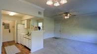 This single-floor residence offers a well-designed open-concept layout that seamlessly connects the kitchen and living area, creating a functional and inviting atmosphere. The kitchen features white cabinetry, a built-in sink, and a peninsula counter with a pass-through opening that looks directly into the main living space, making it ideal for entertaining or everyday convenience. The kitchen is finished with hardwood-style flooring, while the living area transitions to plush wall-to-wall carpeting, adding warmth underfoot. Crown molding runs along the ceilings throughout, lending a clean, polished look to the interiors. The living area is equipped with a ceiling fan with integrated lighting, complementing the elegant chandelier-style light fixture near the kitchen pass-through. A rear entry door provides direct access from the living space, and a window in the kitchen allows for natural light to filter through. The overall color palette is light and neutral, with white trim and soft wall tones that contribute to a bright, airy ambiance. The space is thoughtfully laid out to maximize usability while maintaining an open, comfortable feel throughout.