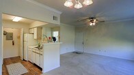 This single-floor residence offers a well-designed open-concept layout that seamlessly connects the kitchen and living area, creating a functional and inviting atmosphere. The kitchen features white cabinetry, a built-in sink, and a peninsula counter with a pass-through opening that looks directly into the main living space, making it ideal for entertaining or everyday convenience. The kitchen is finished with hardwood-style flooring, while the living area transitions to plush wall-to-wall carpeting, adding warmth underfoot. Crown molding runs along the ceilings throughout, lending a clean, polished look to the interiors. The living area is equipped with a ceiling fan with integrated lighting, complementing the elegant chandelier-style light fixture near the kitchen pass-through. A rear entry door provides direct access from the living space, and a window in the kitchen allows for natural light to filter through. The overall color palette is light and neutral, with white trim and soft wall tones that contribute to a bright, airy ambiance. The space is thoughtfully laid out to maximize usability while maintaining an open, comfortable feel throughout.
