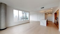This contemporary high-rise residence is defined by clean lines, a neutral palette, and an abundance of natural light. The expansive open-plan living and dining area is anchored by floor-to-ceiling windows that frame sweeping city skyline views, with light oak hardwood flooring running throughout, amplifying the sense of space and brightness.

The kitchen is a standout feature, showcasing rich warm-toned wood cabinetry paired with a striking marble-veined waterfall island topped in white stone. A teal glass tile backsplash adds a refined contrast behind the gas cooktop, while stainless steel appliances and a sculptural multi-arm pendant light complete the contemporary design.

The bedroom is generously proportioned and accesses a well-appointed bathroom featuring a dual-sink vanity with warm wood cabinetry, stone countertops, and a large mirror. A second bathroom offers additional dual undermount sinks set in a dark stone countertop with matching wood cabinetry and large-format tile flooring.

A custom walk-in closet provides extensive built-in shelving, open cubbies, and integrated drawers for organized storage. A dedicated laundry area accommodates a full-size washer and dryer. The overall ambiance is polished, airy, and distinctly urban.