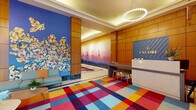 The Encore lobby is a thoughtfully designed, single-floor space that makes an immediate impression through its bold use of color, art, and architectural detail. At its heart is a striking multicolored geometric carpet in vivid shades of red, blue, orange, pink, and teal, which anchors the entire space and sets a lively, energetic tone. Warm, honey-toned wood wall paneling lines the space throughout, lending a refined contrast to the playful flooring below. The front reception desk, branded prominently with the Encore name in gold lettering against a navy blue paneled fascia, serves as the focal point of the primary entry area. A circular pearl-drop chandelier overhead adds an elegant finishing touch to the tray ceiling above. A large-scale floral mural in vivid blues and warm neutrals adorns one wall, while a separate corridor features a gradient ombre wall treatment with decorative lighthouse motifs. The elevator bank is clad in matching wood paneling with polished stainless steel doors, and a sculptural red-and-yellow figure commands attention nearby. Mailboxes, a colorful geometric credenza with a bold red accent wall, and framed photography contribute additional layers of artistic character. Seating areas with contemporary gray sofas and round wood tables provide welcoming gathering spots throughout the lobby.
