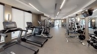 This fitness center offers a spacious, single-floor layout designed to accommodate a full range of workout routines. The space is finished with a durable speckled rubber flooring throughout, providing both comfort and safety during exercise. Recessed linear lighting runs across the clean white ceiling, ensuring the entire facility is brightly and evenly illuminated.

The cardio zone is well-stocked with a row of Cybex treadmills, elliptical trainers, and stair climbers, each equipped with individual display screens and positioned alongside roller blinds that soften the natural light from large windows. The strength training area features a robust selection of Cybex cable and selectorized machines catering to a variety of muscle groups, while a dedicated free weights section includes a full rack of dumbbells, barbells, a weight bench, and a medicine ball station with wall-mounted mirrors that amplify the sense of space.

Large floor-to-ceiling glass partition walls and doors divide sections of the facility, contributing to an open, airy atmosphere while maintaining a polished, modern aesthetic. Neutral taupe-toned walls complement the contemporary design, and wall-mounted televisions are positioned for convenience throughout. The overall environment is clean, organized, and professional, making it a comprehensive facility suited for a wide range of fitness needs.