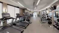 This fitness center offers a spacious, single-floor layout designed to accommodate a full range of workout routines. The space is finished with a durable speckled rubber flooring throughout, providing both comfort and safety during exercise. Recessed linear lighting runs across the clean white ceiling, ensuring the entire facility is brightly and evenly illuminated.

The cardio zone is well-stocked with a row of Cybex treadmills, elliptical trainers, and stair climbers, each equipped with individual display screens and positioned alongside roller blinds that soften the natural light from large windows. The strength training area features a robust selection of Cybex cable and selectorized machines catering to a variety of muscle groups, while a dedicated free weights section includes a full rack of dumbbells, barbells, a weight bench, and a medicine ball station with wall-mounted mirrors that amplify the sense of space.

Large floor-to-ceiling glass partition walls and doors divide sections of the facility, contributing to an open, airy atmosphere while maintaining a polished, modern aesthetic. Neutral taupe-toned walls complement the contemporary design, and wall-mounted televisions are positioned for convenience throughout. The overall environment is clean, organized, and professional, making it a comprehensive facility suited for a wide range of fitness needs.