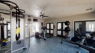 This single-floor fitness center offers a comprehensive workout environment designed to accommodate a wide range of exercise routines. The space is finished with durable, patterned carpet tile flooring in a cool blue-grey tone, and features a textured ceiling with a ceiling fan that aids in air circulation during workouts. Natural light filters in through multiple windows along the walls, creating a bright and energizing atmosphere.

The cardio section includes a treadmill, elliptical, stationary bike, and a rowing machine, providing diverse options for cardiovascular training. A Life Fitness Synergy360 functional trainer serves as a centerpiece for strength and functional fitness, complete with TRX suspension straps. The free weights area is well-stocked with a full rack of kettlebells in varying weights, dumbbells, and medicine balls stored on a dedicated rack. An adjustable workout bench adds further versatility for strength training exercises.

Floor-to-ceiling mirrors line multiple walls, visually expanding the space and allowing users to monitor their form throughout workouts. Black-framed glass doors and wall-mounted televisions add to the polished, modern aesthetic. Safety features such as a fire extinguisher and exit signage are also present. Overall, the space is thoughtfully organized to support both individual training sessions and group fitness activities.