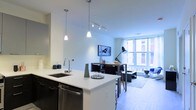 This single-floor residence showcases a thoughtfully designed open-concept layout that seamlessly connects the kitchen, dining, and living areas into one cohesive, functional space. The modern kitchen features dark cabinetry contrasted by crisp white quartz countertops, a stainless steel undermount sink with a pull-down faucet, and a stainless steel dishwasher. Elegant pendant lights hang above the kitchen island, adding a refined touch to the space. A track lighting system overhead provides flexible, adjustable illumination throughout. The dining area is positioned just beyond the kitchen, anchored by a sleek bar-height table and stools, making it an ideal spot for casual meals or working from home. The living area extends toward a generous floor-to-ceiling window that floods the space with natural light and offers views of the surrounding urban environment. A wall-mounted flat-screen television, a contemporary floor lamp, and a neatly arranged seating arrangement complete the living space. Light wood-toned flooring runs throughout, enhancing the bright, airy atmosphere. The overall ambiance is clean, contemporary, and urban — a well-appointed space designed for modern city living.