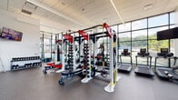 This fitness facility is designed to support a comprehensive workout experience within a spacious, single-level layout. The space is defined architecturally by expansive floor-to-ceiling curtain wall windows that wrap around multiple sides of the room, flooding the interior with abundant natural light and offering unobstructed views of the surrounding landscape and skyline. The soaring ceiling height enhances the open, airy atmosphere throughout.

The strength training area is centrally positioned and anchored by a row of multi-functional cable and power rack systems, including Octagon-branded units equipped with adjustable weight stacks, resistance bands, and TRX suspension attachments. Adjustable benches accompany the racks, providing versatile options for a wide range of strength exercises. A dumbbell rack and additional free weight equipment are visible along the perimeter.

The cardio zone lines the windowed walls, housing a well-curated selection of equipment including treadmills, a StairMaster, rowing machines, and stationary spin bikes, some with integrated digital screens. Wall-mounted televisions are positioned throughout for added convenience.

The flooring throughout is durable rubber gym matting, practical and impact-absorbing. Decorative frosted graphics on the lower window panels add a branded aesthetic touch. The overall ambiance is modern, bright, and professionally appointed.