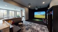 This expansive, single-floor amenity lounge is designed to deliver a premium social and recreational experience. The space is anchored by large floor-to-ceiling windows that flood the interior with natural light and offer open views of the surrounding urban landscape, creating a bright and inviting atmosphere throughout.

The lounge is organized into several distinct activity zones. A dedicated golf simulator room features a full projection screen, real turf hitting mat, and comfortable seating, making it ideal for both practice and entertainment. Adjacent to this, an arcade area houses classic upright gaming cabinets, while a separate zone offers table tennis, air hockey, billiards, and a shuffleboard table — providing a comprehensive range of recreational options.

A central bar-height island with stylish counter seating serves as a natural social hub, complemented by wall-mounted flat-screen televisions positioned throughout the space for easy viewing. Curated artwork and tall potted plants add warmth and visual interest to the neutral gray walls and textured wall coverings.

Throughout the lounge, a bold plaid-patterned carpet in tones of gray, charcoal, and white ties the zones together cohesively. Vibrant terracotta-orange lounge chairs paired with a statement multi-globe chandelier in the seating nook create a striking focal point, balancing comfort with contemporary design.