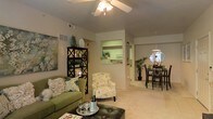 This single-story residence offers an open-concept floor plan that seamlessly connects the living, dining, and kitchen areas into one cohesive, functional space. The living area features plush carpeted flooring and a ceiling fan with integrated lighting, contributing to a comfortable and airy atmosphere. Neutral-toned walls provide a versatile backdrop, complemented by tasteful décor including decorative shelving and botanical artwork.

A pass-through window connects the living space to the kitchen, which is visible in the background and features white cabinetry and standard appliances, creating an accessible and practical cooking environment. The kitchen's green-accented walls add a subtle pop of color that ties into the overall design palette.

Adjacent to the living area, the dining space is defined by an elegant chandelier-style pendant light and accommodates a dark-toned dining set, making it well-suited for both everyday meals and entertaining. The open layout allows natural sightlines across all three areas, enhancing the sense of spaciousness throughout. Overall, the space presents a warm, welcoming ambiance with a cohesive design aesthetic.