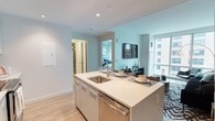 This well-appointed 1-bedroom, 1-bathroom residence is designed with a clean, modern aesthetic throughout. The open-concept layout seamlessly connects the kitchen, dining, and living areas, creating an inviting and functional flow. The kitchen serves as a central hub, anchored by a sleek island with a crisp white countertop, an undermount stainless steel sink, and a stainless steel dishwasher. Light wood-toned cabinetry adds warmth while maintaining a contemporary feel. Soft blue-green accent walls complement the neutral tones, giving the space a calm and airy ambiance. Large windows flood the living area with natural light and offer views of the surrounding cityscape, enhancing the open feel of the space. The living area is comfortably sized and connects naturally to the dining space, which features a modern table and chairs. The bedroom is accessible through a dedicated hallway, offering a sense of privacy and separation from the main living areas. Light hardwood-style flooring runs throughout, tying each area together cohesively. The bathroom, visible from the hallway, continues the clean, modern design language found across the residence. Overall, the space offers a bright, functional, and thoughtfully designed urban living environment.