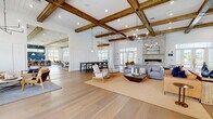 This single-story community clubhouse blends modern farmhouse design with warm, inviting finishes throughout its expansive open-concept floor plan. Wide-plank light hardwood flooring flows seamlessly across every area, unifying the space and enhancing its airy feel. Soaring ceilings adorned with rustic recessed wood box beams serve as a defining architectural feature, complemented by stylish pendant and chandelier lighting that adds warmth and dimension.

The main gathering area is anchored by a floor-to-ceiling stacked stone fireplace with a linear hearth, surrounded by comfortable lounge seating and accent chairs. An adjacent dining area features a long communal table and built-in banquette seating with channeled upholstery, creating an inviting spot for shared meals or casual gatherings. A separate lounge area includes a shuffleboard table, soft seating, and a wall-mounted display screen, ideal for recreation and entertainment.

A well-appointed café-style bar area showcases navy blue glazed subway tile backsplash, white cabinetry, and under-counter refrigerators. The leasing office area offers dedicated workstations with accent wallpaper and elevated pendant lighting. Large glass entry doors and expansive windows throughout flood the space with natural light and provide views of the surrounding community and outdoor pool area.