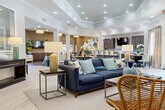 The clubhouse at Cahaba Grandview in Birmingham, Alabama, is a thoughtfully designed single-floor community space that blends sophistication with comfort. The expansive open-plan layout features light hardwood-style flooring throughout much of the space, complemented by tray ceilings with recessed lighting that lend architectural depth and a refined ambiance.

The main lounge area serves as a central gathering point, furnished with navy blue sofas, rattan accent chairs, and layered lighting including a sculptural gold chandelier. A dramatic stacked-stone fireplace wall anchors one section of the lounge, with a mounted television above and an adjacent game room featuring a foosball table. Large windows dressed with neutral drape panels invite generous natural light and outdoor views throughout.

A well-appointed kitchen and dining area features dark wood cabinetry, mosaic tile backsplash, a breakfast bar with bar seating, pendant lighting, and a wall-mounted television — ideal for social gatherings. A separate dining area with a round table provides additional seating. A private leasing office with carpeted flooring, coastal artwork, and a welcoming reception desk completes the space, along with a cozy seating nook featuring shiplap walls and vibrant decorative accents.