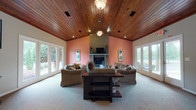 Located in Griffin, Georgia, this single-story property offers a thoughtfully designed layout highlighted by rich architectural details throughout. The centerpiece of the space is a grand living area featuring a soaring vaulted ceiling finished in warm, natural wood planking that lends a distinctive character to the entire room. Walls in a warm terracotta hue frame a full brick fireplace with a classic mantel, creating a welcoming focal point. Large windows and glass doors line the walls on multiple sides, flooding the room with abundant natural light and providing views of the surrounding greenery outdoors. Plush wall-to-wall carpeting adds comfort underfoot across the main living zone. Adjacent to the living area, a pass-through bar counter connects to the kitchen, which features rich wood cabinetry and modern appliances. Pendant lighting above the bar area and recessed lighting throughout complement the wood ceiling aesthetic seamlessly. A dedicated workspace area is also present, equipped with built-in wood cabinetry, ample desk space, and direct exterior access through a glass-paneled door. The consistent use of wood-toned ceilings, neutral carpeting, and warm accent walls creates a cohesive and inviting ambiance throughout the entire property.