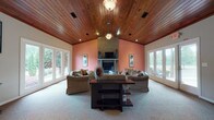 Located in Griffin, Georgia, this single-story property offers a thoughtfully designed layout highlighted by rich architectural details throughout. The centerpiece of the space is a grand living area featuring a soaring vaulted ceiling finished in warm, natural wood planking that lends a distinctive character to the entire room. Walls in a warm terracotta hue frame a full brick fireplace with a classic mantel, creating a welcoming focal point. Large windows and glass doors line the walls on multiple sides, flooding the room with abundant natural light and providing views of the surrounding greenery outdoors. Plush wall-to-wall carpeting adds comfort underfoot across the main living zone. Adjacent to the living area, a pass-through bar counter connects to the kitchen, which features rich wood cabinetry and modern appliances. Pendant lighting above the bar area and recessed lighting throughout complement the wood ceiling aesthetic seamlessly. A dedicated workspace area is also present, equipped with built-in wood cabinetry, ample desk space, and direct exterior access through a glass-paneled door. The consistent use of wood-toned ceilings, neutral carpeting, and warm accent walls creates a cohesive and inviting ambiance throughout the entire property.