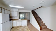 This two-story residence offers a practical and comfortable layout spread across two levels. The main floor features an open-plan kitchen and living area, where the kitchen is equipped with white cabinetry, a countertop with an integrated sink, a dishwasher, and tile flooring that adds a clean, durable foundation to the space. The kitchen flows seamlessly into the adjoining living area, which features carpeted flooring, a ceiling fan, and a sliding glass door that invites natural light and provides access to the outdoors. The transition between the two flooring materials creates a clear, functional distinction between the kitchen and living zones. A staircase with a warm wood banister and black metal balusters connects the main level to the upper floor, adding a classic design element to the interior. The white walls and neutral tones throughout create a bright, airy ambiance, while the ceiling fixtures provide ample lighting. The overall layout is well-organized, making efficient use of the available space across both levels.