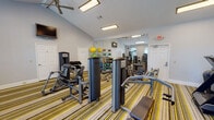 This fitness center is a comprehensively outfitted exercise facility designed to support a full range of workout routines. The space features vaulted ceilings that lend an open, airy feel, complemented by ceiling fans that promote air circulation throughout. Large arched windows along one wall invite abundant natural light and offer views of the surrounding exterior, creating an energizing atmosphere for workouts.

The floor is covered in a bold, striped carpet in tones of yellow, green, and gray, adding a vibrant visual character to the space. A full-length wall mirror runs along one side of the room, effectively doubling the perceived size of the facility while allowing users to monitor their form during exercise.

The strength training area is well-appointed with multiple cable and resistance machines, a dumbbell rack stocked with a range of weights, and various seated press and pull machines. The cardio section features treadmills, an elliptical trainer, a stationary bike, and a stair climber, catering to a variety of aerobic preferences. A medicine ball rack with an assortment of weighted balls adds functional training options to the mix.

A wall-mounted flat-screen television provides entertainment, while flush panel lighting ensures the space remains well-lit at all hours. The clean, light gray walls and organized layout contribute to a focused and motivating workout environment.