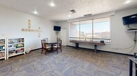 Located at 1100 Penn Center Blvd in Pittsburgh, PA, this single-floor game room is a well-appointed recreational space designed for leisure and social engagement. The room features patterned carpet throughout, adding a dynamic visual element to the overall design, while white walls and recessed lighting create a bright, welcoming atmosphere.

The space is anchored by a full-size shuffleboard table positioned along a wall of large windows that flood the room with natural light and offer expansive views of the surrounding Pittsburgh skyline and rolling landscape. A round wooden table with chairs provides an ideal setting for card games or casual conversation, complemented by wall-mounted flat-screen televisions for entertainment.

A dedicated area houses a classic mini arcade cabinet, accompanied by a table and seating for additional gaming options. A white bookshelf stocked with an assortment of board games lines one wall, ensuring a variety of entertainment choices. Decorative video game-themed framed artwork and Scrabble-tile wall lettering add personality and a playful character to the space.

The game room is accessible through a glass-paneled door and operates during designated hours, making it a communal hub for recreation. The combination of games, natural light, and city views makes this space both functional and inviting.