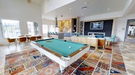 This thoughtfully designed amenity lounge combines recreation, socializing, and relaxation into one cohesive, open-plan space. Soaring ceilings lend an airy, expansive feel, while large windows along the perimeter allow natural light to flood the interior. At the heart of the room sits a sleek, contemporary pool table with a teal felt surface and crisp white frame, anchored atop a vibrant, patchwork-style area rug that adds warmth and character to the polished floor.

The kitchenette and bar area features gray cabinetry, a countertop workspace, and bar-height seating with blue upholstered stools, all set against a striking geometric gold-toned accent wall that serves as a bold architectural focal point. Pendant lighting above the bar enhances the modern aesthetic.

Opposite the kitchenette, a dedicated media and lounge zone offers built-in shelving, a wall-mounted flat-screen television, and comfortable seating centered around a fireplace framed by richly patterned navy wallcovering. Natural wood-toned furniture and soft textiles create a welcoming, relaxed atmosphere. A casual dining nook with additional seating is tucked near the windows, rounding out this multifunctional and well-appointed communal space.