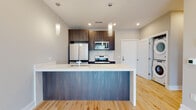 This contemporary single-floor residence showcases a thoughtfully designed open-concept layout with high-end finishes throughout. The kitchen serves as a central focal point, featuring dark wood cabinetry, a full suite of stainless steel appliances including a refrigerator, electric range, and built-in microwave, all complemented by a striking large-format island with a white marbled quartz countertop and a wood-paneled base. Elegant cylindrical pendant lights hang above the island, adding a refined touch to the space. Light-toned hardwood flooring extends seamlessly throughout the unit, enhancing the bright, airy ambiance. The living area is anchored by a built-in linear fireplace with a dedicated media niche above, creating a sleek and functional entertainment wall. A sliding glass door in the bedroom area fills the room with natural light and offers outdoor views, further contributing to the open feel of the space. A stacked washer and dryer unit is conveniently integrated into a dedicated nook near the kitchen. The bathroom is finished with floor-to-ceiling marble-look tile and features a frameless glass-enclosed walk-in shower with a multi-function chrome shower system, paired with a floating wood-toned vanity for a clean, modern aesthetic.