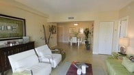 This single-floor residence presents an open-concept design that seamlessly connects the living and dining areas, creating a cohesive and functional living environment. The interior is finished in warm, creamy neutral tones across both the walls and tile flooring, lending a clean and consistent aesthetic throughout the space.

The living area is furnished with contemporary white leather seating, including a sofa and accent chair with chrome frames, arranged around a low coffee table atop a rich burgundy area rug. Decorative bamboo stalks and framed wall art add visual interest and a subtle natural element to the room.

The dining area, visible through an open archway, features a round white dining table surrounded by matching white chairs, illuminated by a pendant light fixture above. Lush potted tropical plants flank the dining space, enhancing the airy, resort-like atmosphere. Framed artwork and a window bringing in natural light complete the dining alcove.

The tile flooring runs continuously throughout, contributing to the spacious, uninterrupted feel of the layout. The overall ambiance is bright, modern, and inviting, with a clean design sensibility suited to comfortable everyday living.