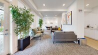 Located at 46120 Thomas Rd in Chilliwack, BC, this thoughtfully designed single-floor amenity space offers a welcoming environment for residents to gather, relax, and connect. The open-concept layout is anchored by a spacious living area furnished with contemporary grey sofas, accent chairs, and warm yellow throw pillows, all set atop textured carpet flooring. Tall bamboo plants serve as natural dividers, adding greenery and a sense of calm throughout the space.

Recessed ceiling lighting illuminates the crisp white walls, which are accented by curated artwork, including an abstract colorful canvas that adds a vibrant touch. Large roller-shade windows invite natural light and provide views to the exterior entry.

A dedicated dining nook features a round wood table surrounded by modern black-and-walnut chairs, complemented by a bar-height counter with stools — ideal for casual meals or informal gatherings. An additional lounge nook offers a cozier setting with a round table, matching seating, a well-stocked bookshelf, and a large wall-mounted display screen, creating an inviting reading and games area.

Structural columns are seamlessly integrated into the design, and the overall aesthetic balances modern furnishings with warm, natural accents, resulting in a polished yet comfortable communal environment.