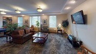 This single-floor communal amenity space is thoughtfully designed to offer residents a variety of recreational and social opportunities within a warm, inviting atmosphere. The layout flows seamlessly across several distinct activity zones, all unified by patterned carpet flooring featuring an elegant floral motif and tray ceilings with recessed lighting throughout.

The main lounge area serves as the heart of the space, furnished with plush, dark-upholstered sofas and armchairs arranged around a glass-topped coffee table. Large windows along one wall invite an abundance of natural light and provide pleasant views of the surrounding greenery, while a wall-mounted flat-screen television adds a modern entertainment option. Lush potted plants are positioned throughout, adding a refreshing, natural element to the interior.

Adjacent to the lounge is a dedicated billiards area, centered around a full-sized pool table with a vibrant blue felt surface. Built-in bookshelves flank a classic wood-mantel fireplace nearby, creating a cozy library nook complete with upholstered armchairs — an ideal retreat for reading or quiet conversation.

Connecting corridors feature additional seating, including a charming wooden rocking chair, and lead to further areas of the facility. Bulletin boards are thoughtfully placed throughout, supporting community communication. The overall ambiance is comfortable, residential, and community-focused.