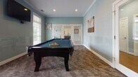 This single-floor community amenity space is thoughtfully designed to offer residents a variety of recreational and functional areas, all unified by a cohesive soft blue color palette, white crown molding, and patterned carpet flooring throughout.

The game room serves as a centerpiece of the space, featuring a full-size black pool table set beneath recessed lighting. Multiple windows dressed with white blinds allow natural light to filter in, creating a bright and inviting atmosphere, while framed wall art adds a decorative touch.

Adjacent to the game room is a comfortable lounge and seating area furnished with dark wood tables and mixed metal and wood chairs. Glass-paneled doors and windows provide views of the outdoors, enhancing the open feel of the space.

A dedicated business center offers multiple computer workstations arranged along a built-in desk, complemented by a wall-mounted flat-screen television and additional seating — ideal for productivity or presentations.

The café bar area features rich dark wood cabinetry, granite-style countertops, and under-cabinet lighting. Equipped with a coffee maker and condiment station, it serves as a convenient refreshment hub for the space.

Together, these areas create a versatile, well-equipped environment suited for socializing, working, and recreation.