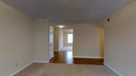 This single-floor residence offers a straightforward and functional layout with a clean, understated aesthetic. The interior is finished in soft, warm neutral tones throughout, creating a cohesive and welcoming atmosphere across all rooms. The ceilings feature a classic textured popcorn finish, adding subtle visual character overhead. Flooring transitions between plush carpet in the main living area and rich hardwood in the connecting hallway and adjacent room, providing both comfort and practicality. White baseboards run consistently along the walls, lending a crisp, finished look to each space. The layout flows openly from the main living area through a wide passageway into a secondary room, which benefits from natural light filtering in through a window. A doorway off the main area leads to additional spaces, suggesting a well-connected floor plan designed for ease of movement. Flush-mount ceiling lighting provides steady, even illumination throughout. Overall, the space presents a modest yet functional interior with classic residential finishes, offering a neutral canvas well-suited for a variety of furnishing styles and personal touches.