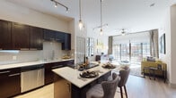 This single-floor residence showcases a thoughtfully designed open-concept layout that seamlessly connects the kitchen, dining, and living areas. The kitchen is a standout feature, equipped with dark espresso cabinetry extending to the ceiling, stainless steel appliances including a French door refrigerator and dishwasher, a textured tile backsplash, and light quartz countertops. A central island with bar seating serves as both a prep space and casual dining area, illuminated by modern glass pendant lights and complemented by a track lighting system overhead.

The living area benefits from generous natural light streaming through expansive floor-to-ceiling windows and a sliding glass door that opens to an outdoor space. A ceiling fan adds to the comfort of this bright, airy room. Light wood-style flooring runs throughout the main living spaces, enhancing the contemporary aesthetic.

The bedroom is a calm retreat featuring multiple windows with blinds that allow natural light while maintaining privacy, a tray ceiling detail, ceiling fan, and wood-style flooring. A hallway leads to a dedicated walk-in closet with built-in shelving, providing substantial storage. The bathroom features tile flooring and dark cabinetry, consistent with the unit's cohesive modern design language throughout.