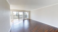 This single-floor space presents a clean, well-maintained interior defined by rich, dark hardwood flooring that extends throughout the open living area. The room is generously proportioned, offering a spacious feel well-suited for a variety of furniture arrangements. Crisp white walls and white crown moulding frame the interior, contributing to a polished and cohesive aesthetic. Natural light floods the space through a large sliding glass door and an adjacent window, both positioned along one wall to maximize brightness throughout the day. The sliding glass door provides direct access to an outdoor balcony enclosed by a dark metal railing, which overlooks a lush, tree-lined setting — offering a pleasant sense of privacy and greenery. The overall ambiance is calm and airy, with the combination of natural light, neutral tones, and warm wood flooring creating an inviting atmosphere. The single-floor layout ensures seamless flow and accessibility throughout the space.