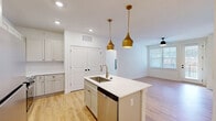 Located at 382 Red Bud Cir in Hendersonville, NC, this well-designed single-floor home offers 2 bedrooms and 2 bathrooms with a clean, modern aesthetic throughout. The open-concept kitchen and living area serves as the heart of the home, featuring a central island with a stainless steel sink, sleek white quartz countertops, white shaker cabinetry with gold hardware, and a full suite of stainless steel appliances including a range, microwave, and refrigerator. A white subway tile backsplash and a pair of striking gold pendant lights add a refined, contemporary touch. Light hardwood flooring flows throughout the main living areas, complementing the bright, white walls and recessed lighting. The living room receives ample natural light through large windows with blinds and includes a ceiling fan and a private exterior door. Both bedrooms are carpeted and feature ceiling fans, while the primary bedroom benefits from a spacious walk-in closet with full wire shelving systems. The bathrooms maintain the home's clean design language, with light wood-tone flooring, wide vanities with ample cabinetry, and crisp white finishes. A dedicated hallway connects the rooms efficiently, completing this thoughtfully laid-out home.