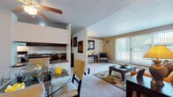 This single-floor residence at 26101 Country Club Blvd in North Olmsted, Ohio, offers a well-organized layout connecting living, dining, kitchen, and sleeping areas in a cohesive flow. The living room is a comfortable, carpeted space anchored by a large sliding glass door with vertical blinds that invite generous natural light and provide views of the surrounding outdoor area. A wall-mounted air conditioning unit and baseboard heating ensure year-round comfort. The dining area, positioned adjacent to the living room and kitchen pass-through, features a ceiling fan overhead and tile flooring at the entry transition. The galley-style kitchen is equipped with dark wood cabinetry, granite-look countertops, a full-size refrigerator, and an electric range with overhead ventilation. The primary bedroom is generously sized with plush carpeting, a walk-in closet, and direct access to an en-suite bathroom. The second bedroom also offers carpet, a standard closet with bi-fold doors, and a window with outdoor light. Two full bathrooms are present — one with a tub/shower combination and white tile flooring, the other with a vanity and mirrored medicine cabinet. The entryway features tile flooring, providing a clean transition into the carpeted main living spaces throughout this well-appointed home.