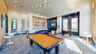 Located at 3445 Bluebonnet Blvd in Baton Rouge, Louisiana, this single-floor community lounge is a thoughtfully designed social space that blends bold aesthetics with functional amenities. The room is immediately distinguished by its dramatic double-height ceilings and striking color palette, pairing deep navy blue accent walls with a vibrant amber-orange feature wall adorned with decorative sconce lighting and framed artwork. Tall, floor-to-ceiling windows flood the space with generous natural light while offering exterior views of the surrounding community. The patterned carpet in blue and white anchors the room and ties together the eclectic furnishings. A central seating area features a plush navy sectional sofa and warm cognac-toned accent chairs arranged around a coffee table, creating an inviting space for relaxed gatherings. A wall-mounted flat-screen television sits within a dark navy built-in nook, flanked by open shelving and a credenza below. Recreation is a highlight, with both a full-size billiard table and a foosball table providing entertainment options. High-top bistro tables and modern bar stools positioned near the windows offer casual seating alternatives. An artistic textured wall panel in gold and white adds a sculptural focal point, complemented by a contemporary branching chandelier overhead. The overall ambiance is energetic, modern, and welcoming.