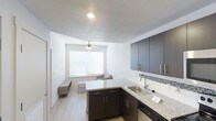 This single-floor space showcases an open-concept layout that seamlessly connects the kitchen and living area, creating a cohesive and functional environment. The kitchen is outfitted with dark-toned cabinetry featuring brushed nickel hardware, a granite-style countertop, and a stainless steel undermount sink with a contemporary gooseneck faucet. A decorative mosaic tile backsplash adds a refined accent between the upper and lower cabinets, while a dishwasher provides added convenience. The countertop extends into a peninsula configuration, offering additional workspace and a natural transition into the living area. Light wood-tone laminate flooring runs throughout, contributing to a clean, modern aesthetic. The living area benefits from a centered window fitted with horizontal blinds, allowing for adjustable natural light. A ceiling fan with an integrated light fixture provides both airflow and ambient lighting. The walls are finished in crisp white, complemented by a textured ceiling treatment. The overall ambiance is bright, contemporary, and efficiently designed, making practical use of the available space.