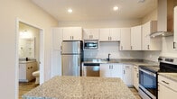 Located at 300 Hickory Trace Ln in Garner, NC, this single-floor residence offers a clean, modern design with thoughtful finishes throughout. The kitchen is a standout feature, equipped with stainless steel appliances including a refrigerator, electric range, built-in microwave, and dishwasher. White shaker-style cabinetry with black hardware lines the walls, complemented by granite countertops and a central island that provides additional prep and storage space. A sleek wall-mounted range hood adds a contemporary touch above the range. Light wood-look flooring runs through the main living areas, creating a cohesive and easy-to-maintain aesthetic. The living area is open and airy, featuring a ceiling fan, neutral warm-toned walls, and a glass door that invites natural light while offering outdoor access. The bedroom is carpeted for comfort, with multiple windows dressed in white blinds that frame views of surrounding greenery and allow ample natural light. A pantry closet is conveniently situated near the kitchen. The bathroom features a combined tub and shower unit with tiled surround, a granite-topped vanity with white cabinetry, and wood-look flooring that ties the space together cohesively. The overall design emphasizes a bright, neutral palette and practical, modern living.