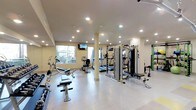 This fitness center offers a comprehensive workout environment designed to accommodate a variety of exercise routines. The space is bathed in bright, even illumination from recessed ceiling lights complemented by natural light streaming through large windows, creating an open and energizing atmosphere. The light-toned walls and pale flooring further enhance the sense of spaciousness throughout the facility. A central support column divides the floor plan subtly without interrupting the overall flow of the space. The cardio section features treadmills, a stationary upright bike, and a recumbent bike, providing ample options for aerobic training. The strength training area is anchored by a multi-station cable machine, offering a versatile range of resistance exercises, while a dedicated free weights section with a bench press station rounds out the muscle-building options. A wall-mounted television is positioned for convenient viewing during workouts. The facility appears well-maintained and thoughtfully arranged to maximize usability, making efficient use of its single-floor layout to serve a broad range of fitness goals.
