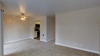This single-floor space offers a straightforward, functional layout designed to maximize usability. The living area is spacious and open, finished with wall-to-wall carpet in a neutral beige tone that adds warmth underfoot. The walls are painted in a soft gray-beige hue, contributing to a clean and understated aesthetic throughout. A ceiling fan with a light fixture provides both overhead illumination and air circulation to the main living space.

A pass-through opening connects the living area directly to the kitchen, which features white cabinetry providing ample storage. The kitchen is equipped with black appliances, including a microwave and a range, and transitions to hard flooring, creating a practical distinction between the two areas. A small wall-mounted vent unit is also visible in the living space, suggesting a built-in climate control feature.

The clean lines, minimal detailing, and neutral color palette throughout give the space a versatile quality, making it easy to personalize with furnishings and décor. The open layout between the living area and kitchen supports a connected, easy-flowing floor plan suited for everyday living.