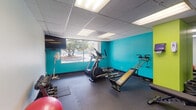 This fitness center offers a practical and energizing workout environment designed to support a range of exercise routines. The space is immediately distinguished by its bold color scheme, featuring vivid teal and lime green accent walls that create a motivating atmosphere against a neutral drop-tile ceiling fitted with recessed fluorescent lighting. Durable dark rubber flooring with interlocking mat sections covers the entire workout area, providing both safety and comfort underfoot. A large window along one wall allows natural light to filter in, offering views of surrounding greenery and contributing to an open, airy feel. A full-length wall mirror on one side of the room visually expands the space and allows users to monitor their form during exercise. Equipment includes an elliptical trainer and a weight bench with an attached resistance system for strength training. A free weight station features a tiered dumbbell rack with multiple weight options, accompanied by a stability ball and a body weight scale. A wall-mounted flat-screen television provides entertainment during workouts, while a standing fan ensures air circulation throughout the room. A rack of medicine balls rounds out the strength training offerings. The overall layout is efficiently organized, making the most of the available footprint while keeping the space accessible and easy to navigate.