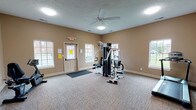 The fitness center at Stonebridge Apartments is a single-story amenity space designed to support residents' health and wellness routines. The interior is finished with neutral tan walls and durable patterned carpeting throughout, creating a clean and functional environment. Flush-mount ceiling lights and a ceiling fan provide consistent illumination and air circulation across the main exercise area.

The space is equipped with a multi-station cable weight machine that supports a variety of strength-training exercises, a treadmill for cardiovascular workouts, and a recumbent stationary bike housed in a dedicated corner area. An open, carpeted flex room connects to the main workout area, offering additional space that could accommodate stretching, floor exercises, or light activities.

Multiple windows line the walls, allowing natural light to fill the interior and providing views of the surrounding community grounds. A keyed exterior door with a glass panel offers direct outdoor access, adding convenience for residents. Safety features include clearly marked emergency exit signage and a security camera. The overall layout is open and uncluttered, allowing for comfortable movement between equipment. The facility presents a practical and well-maintained setting for everyday fitness use.