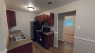 This single-story unit offers a practical and cohesive layout across its main living areas, bedrooms, and bathroom. The kitchen is well-equipped, featuring rich dark wood cabinetry, granite-style countertops, and black appliances including a refrigerator, electric range, and dishwasher. A double sink and ample upper and lower cabinet storage add to its functionality. Dark wood-style laminate flooring runs throughout the kitchen and living areas, providing a clean, durable surface. An open passageway connects the kitchen to the adjacent living space, which features multiple windows fitted with horizontal blinds, allowing natural light to brighten the interior.

The bedrooms are carpeted in neutral tones and include windows with blinds, offering natural light and a comfortable feel. Each room provides a clean, versatile space suitable for various furniture configurations. Select rooms include a chair rail detail along the walls, adding a subtle architectural touch.

The bathroom is compact and efficiently designed, featuring a white vanity with under-cabinet storage, a wall-mounted mirror, a full tub-shower combo, and a toilet. Textured ceilings are consistent throughout the unit, complementing the neutral gray wall palette that unifies the overall aesthetic.