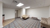 This single-floor space is a well-appointed, self-contained room designed to serve a variety of functional purposes, whether as a lounge, waiting area, or gathering space. The room is finished with a distinctive geometric-patterned carpet in muted tones of brown, blue, and cream, adding a layer of visual interest to the otherwise neutral interior. The walls are painted in a clean, off-white tone that contributes to a calm and professional atmosphere throughout.

The drop ceiling is fitted with large recessed panel lights that provide bright, even illumination across the entire room. A structural column near the center of the space subtly divides the room into distinct zones without closing off the open feel. A set of heavy-duty double doors with push-bar hardware serves as the main entry point, suggesting this space is part of a larger facility.

Along one side of the room, rich dark-wood cabinetry with dark granite-style countertops provides built-in storage and workspace, lending a touch of warmth and practicality to the space. The seating area features a tufted cream-colored sofa and matching chair alongside a simple wooden bench, creating a welcoming lounge environment. A secondary interior door offers additional access. The overall design is clean, functional, and well-maintained, suited for professional or communal use.