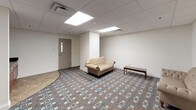 This single-floor space is a well-appointed, self-contained room designed to serve a variety of functional purposes, whether as a lounge, waiting area, or gathering space. The room is finished with a distinctive geometric-patterned carpet in muted tones of brown, blue, and cream, adding a layer of visual interest to the otherwise neutral interior. The walls are painted in a clean, off-white tone that contributes to a calm and professional atmosphere throughout.

The drop ceiling is fitted with large recessed panel lights that provide bright, even illumination across the entire room. A structural column near the center of the space subtly divides the room into distinct zones without closing off the open feel. A set of heavy-duty double doors with push-bar hardware serves as the main entry point, suggesting this space is part of a larger facility.

Along one side of the room, rich dark-wood cabinetry with dark granite-style countertops provides built-in storage and workspace, lending a touch of warmth and practicality to the space. The seating area features a tufted cream-colored sofa and matching chair alongside a simple wooden bench, creating a welcoming lounge environment. A secondary interior door offers additional access. The overall design is clean, functional, and well-maintained, suited for professional or communal use.